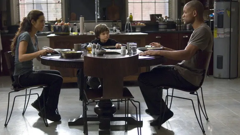 Robert Neville seated at a dining table with his wife and young daughter in I Am Legend