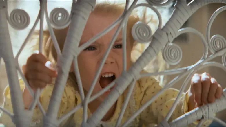 A child gripping the bars of a white bed frame in a bedroom